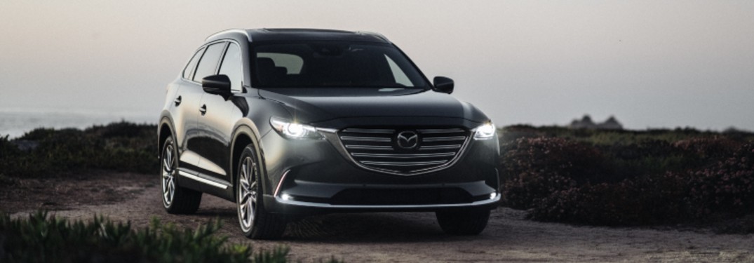 A black 2020 Mazda CX-9 with its lights on at dusk driving down a dirt road.