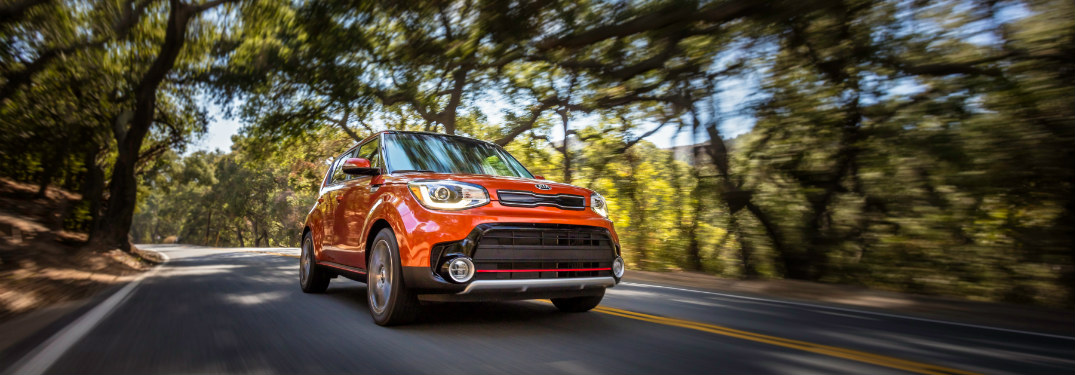 front view of orange 2018 kia soul driving on road surrounded by trees