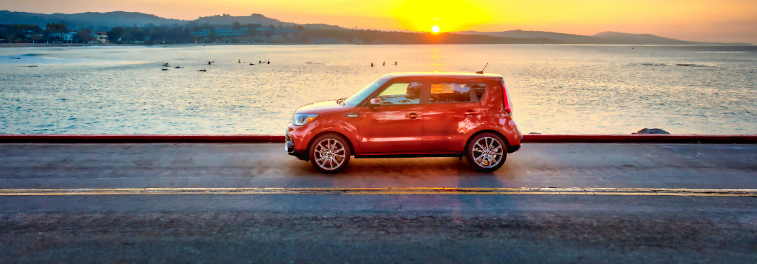 side view of orange 2019 kia soul driving down road with ocean and sun behind it