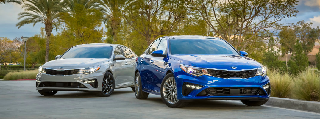 2019 Kia Optima sedan new york international auto show models blue and gray parked on conrete in front of lush tropical green plants