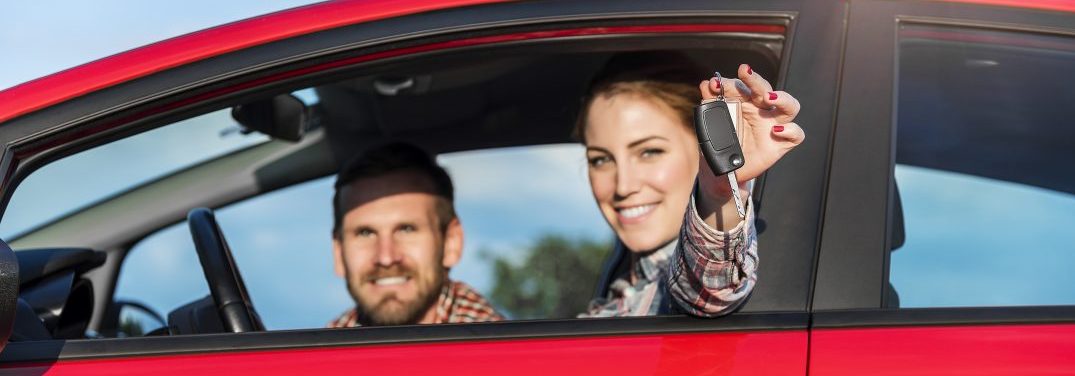 Woman and man sitting in a red car with the woman sitting in the driver seat holding out the keys