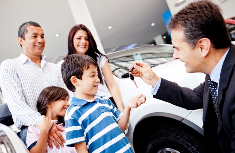 a salesman handing off the keys for a new car purchase at a dealership to the son of a family
