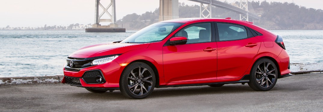 2020 Honda Civic Hatchback exterior side shot with red paint color parked on a beach by the sea with a metal bridge behind it
