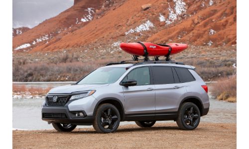 2021 Honda Passport exterior shot with Lunar Silver Metallic paint color parked on a beach near water and mountains with a red kayak on its roof rails