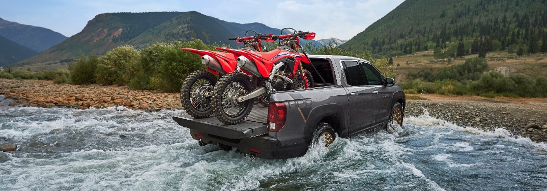 2021 Honda Ridgeline with HPD Package exterior rear shot with motorbikes in its truck bed driving out of water onto a beach of rocks and gravel
