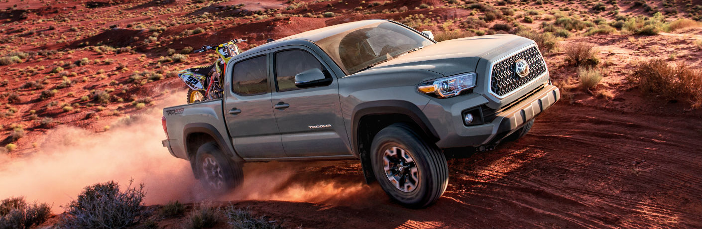 Exterior view of a silver 2018 Toyota Tacoma driving over a desert hill with an ATV in the truck bed