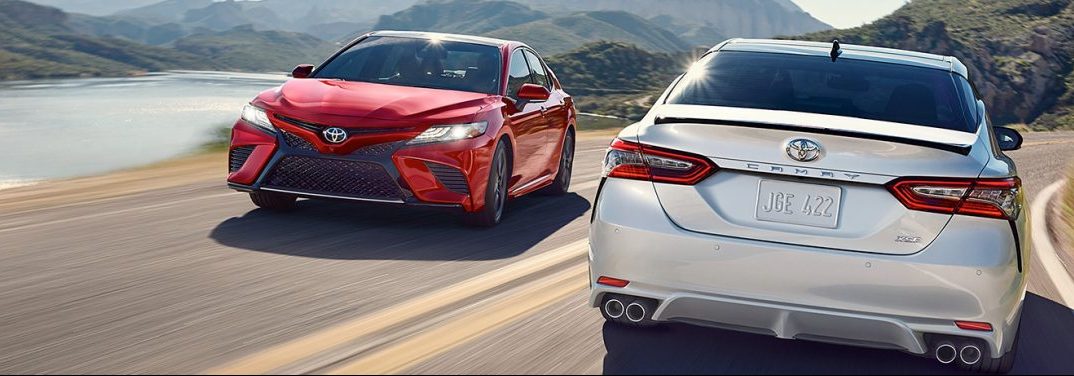 Two 2019 Toyota Camry vehicles drive past each other on a highway. A red model drives towards the camera while a silver drive away, revealing its rear.