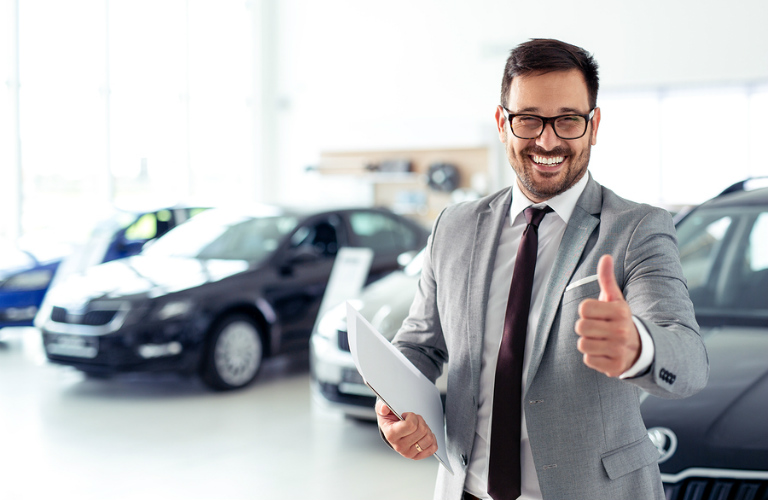 A grinning car salesman gives the thumbs up to the viewer.