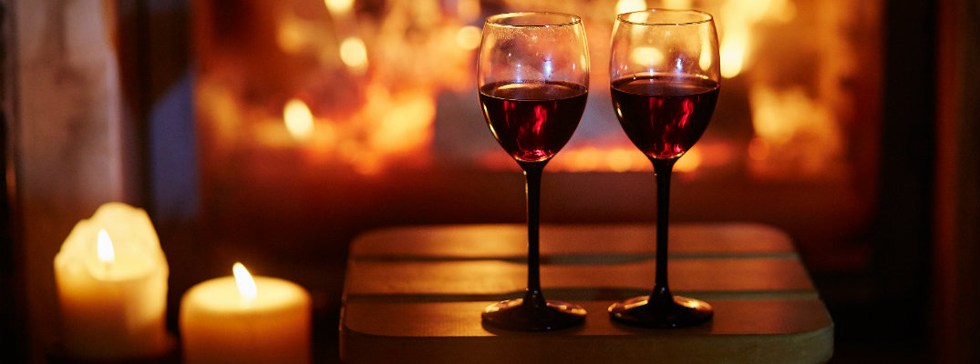 a couple of glasses of red wine set on a stool near lit candles and a fireplace for a romantic Valentine's Day dinner