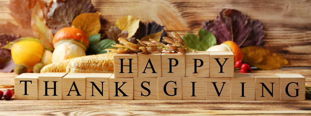 Happy Thanksgiving written out in wooden blocks near a cornucopia of leaves, nuts, corn, vegetables, and other autumn and fall assortments