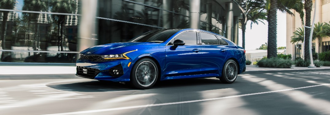 2021 Kia K5 GT exterior shot with blue paint color driving past a hotel surrounded by palm trees