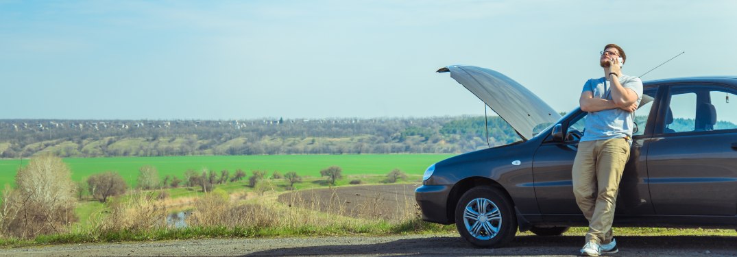 A driver standing next to a broken-down car calling for assistance on a rural road