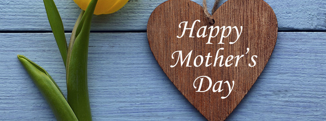 happy mother's day message carved on a wooden heart next to a yellow tulip flower in front of a blue painted house in the grass