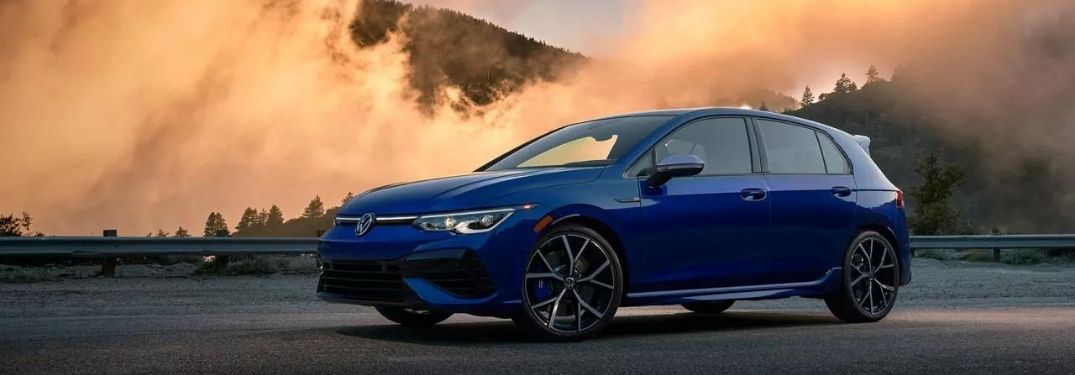 A blue 2022 Volkswagen Golf R parked at an empty space with smoke billowing from the background.