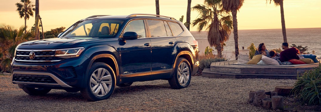 The front and side view of a blue 2021 Volkswagen Atlas parked in front of a couple laying and watching the sunset on a beach.
