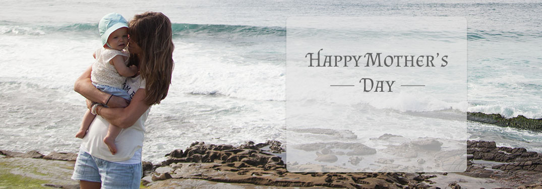 Mother holding baby in front of ocean on overcast day with Mother's Day text
