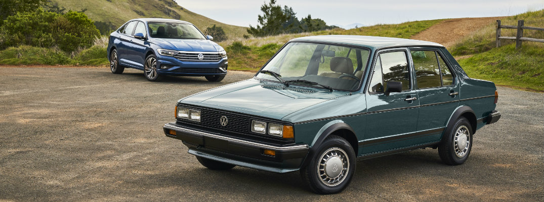 1982 Volkswagen Jetta parked in front of a 2019 Volkswagen Jetta with both models parked on a rocky dirt plain next to grassy hills and a wooden farm fence