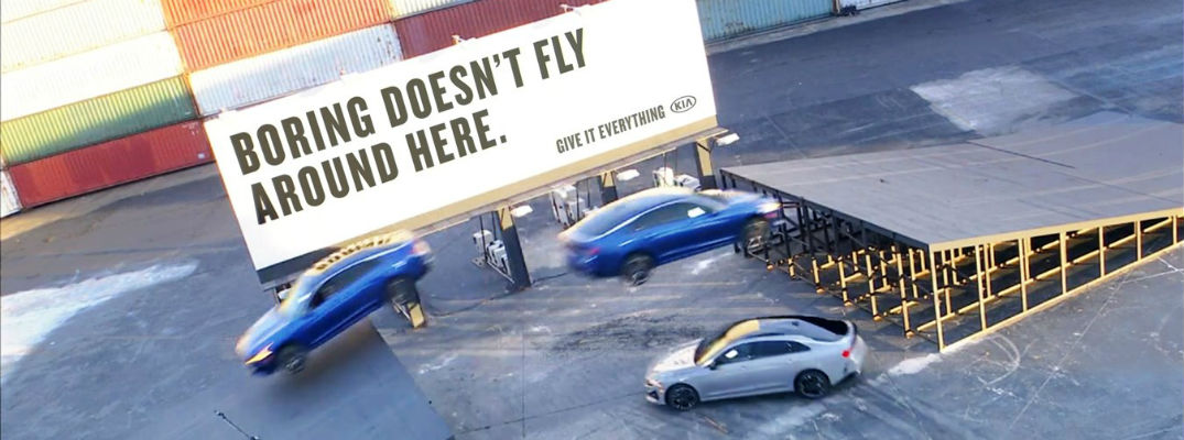 Three Kia K5 vehicles performing Triple Threat Stunt with "Boring Doesn't Fly Around Here" sign