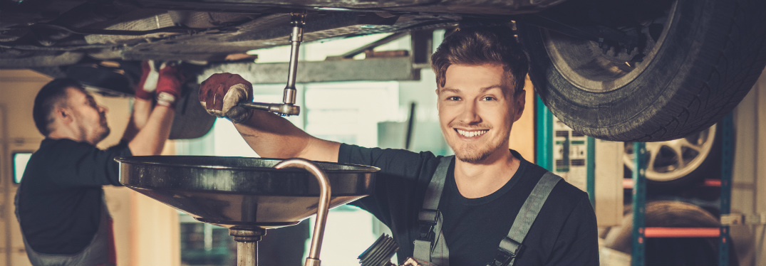 A grinning mechanic performs an unknown procedure on a raised vehicle.