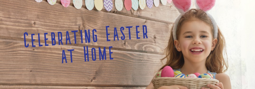 A picture of a little girl with bunny ears indoors smiling with the caption along the wooden wall saying "Celebrating Easter at Home".