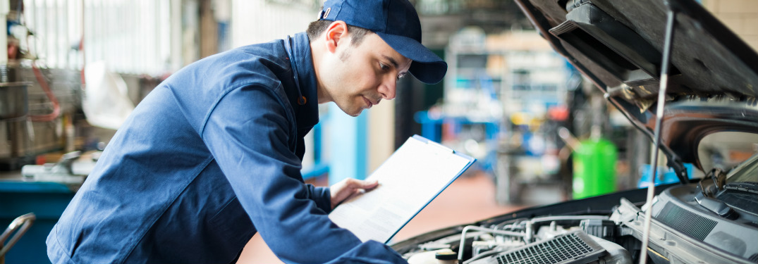 professional mechanic leaning over open hood of car holding clipboard