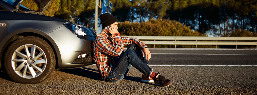 A stock photo of a young man sitting in front of his broken down car, calling for help.