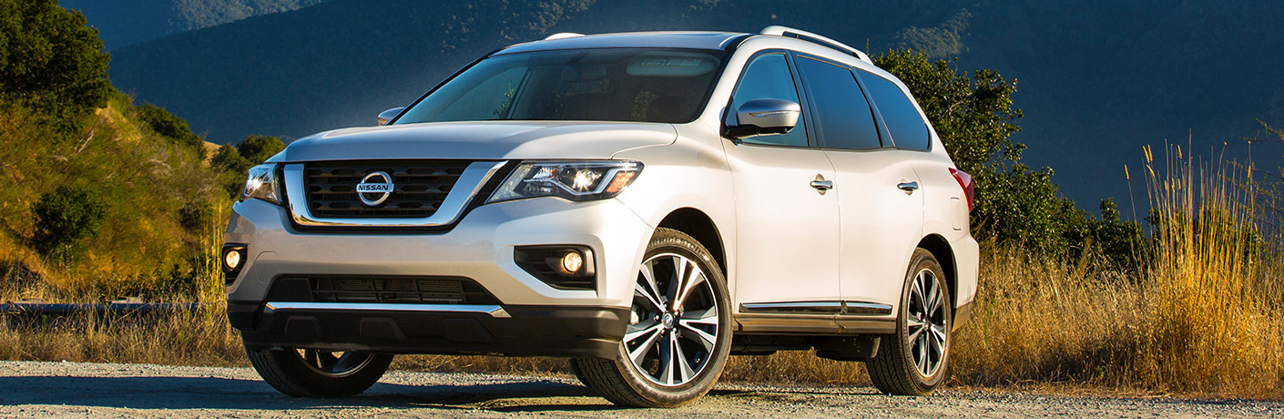 Exterior view of a white 2018 Nissan Pathfinder parked on a highway with trees and tall grass in the background
