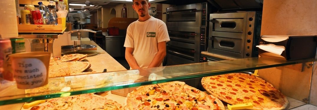 A pizza store with the person at the counter offering a wide variety of options