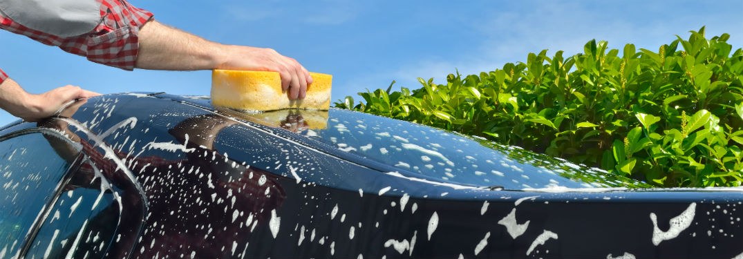 Person wearing a grey shirt with red and white checked cuffs holding a sponge washing their soapy car