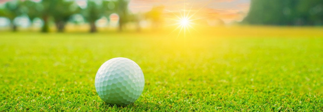 Close up of a golf ball sitting on a golf course with trees and the sun in the background