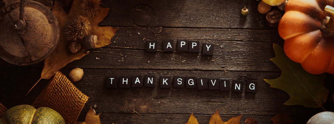 Happy Thanksgiving written out on small tiles on a wooden plank picnic table surrounded by leaves, nuts, and a pumpkin gourd