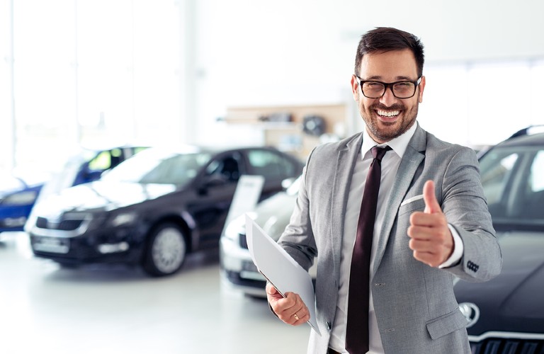 A man in glasses smiling with his thumbs up inside a car dealership.