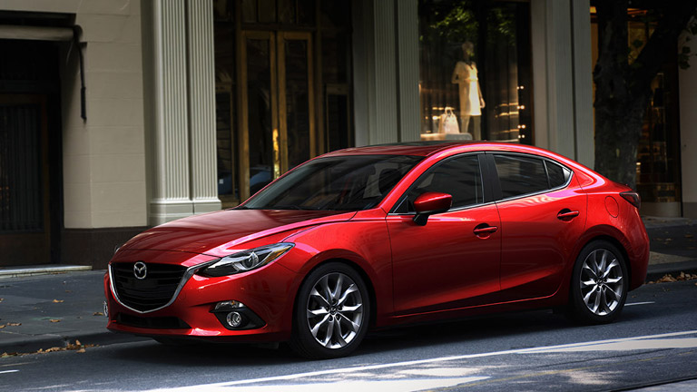 The side view of a red 2015 Mazda3 parked in a city.