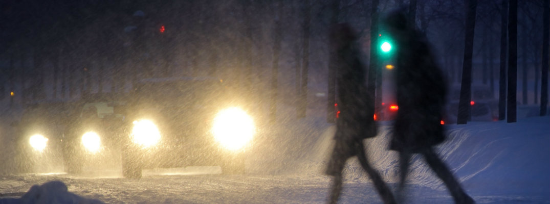2 people crossing the road through snow in front of a line of cars with their lights on
